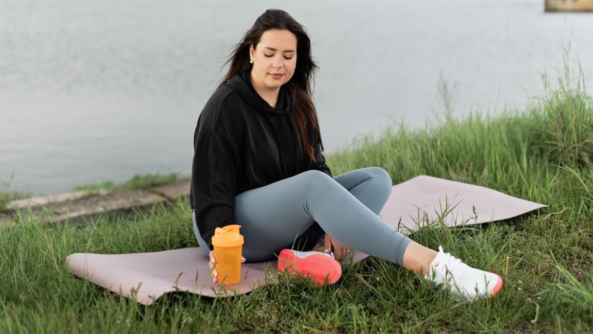 Woman enjoying a peaceful moment by the lake with a yoga mat and water bottle, promoting wellness.
